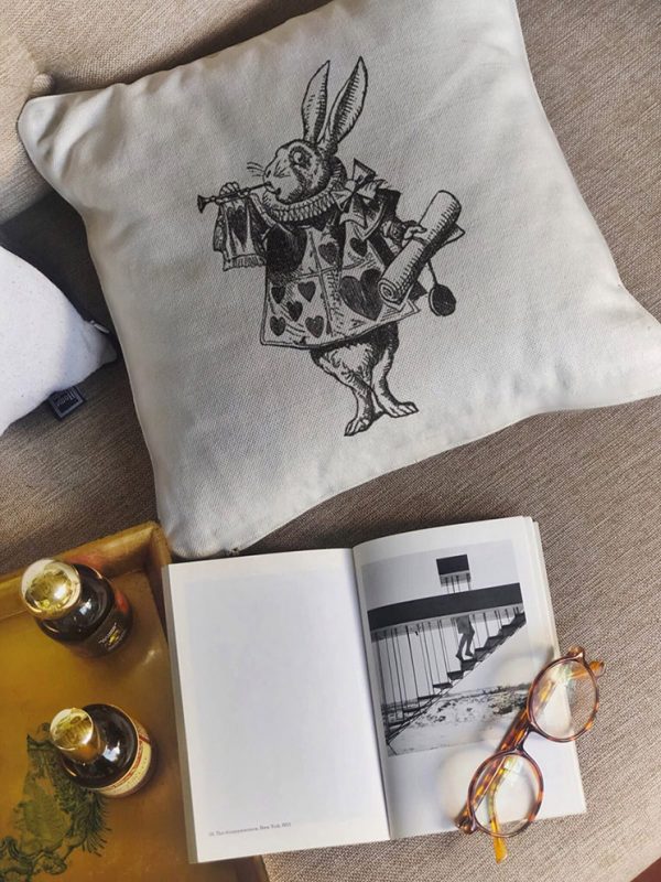 nivens beige cushion made of 100% cotton, messenger rabbit design by Alice in Wonderland on a beige sofa with a book.