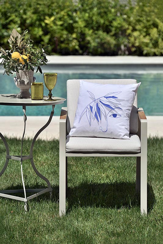 vigilia I Λευκό μαξιλάρι από 100% βαμβάκι, με ιδιαίτερο σχέδιο μπλε πρόσωπο και κλαδί ελιάς πάνω σε καρέκλα κήπου μπροστά από πισίνα.