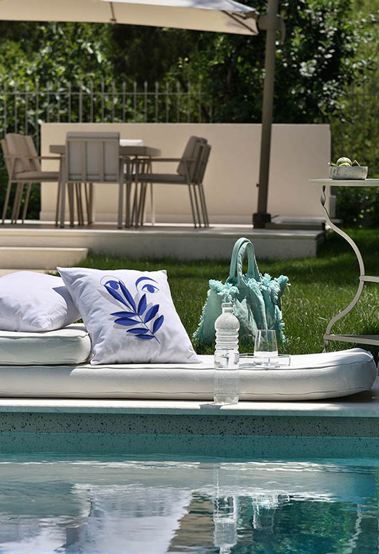 vigilia II Λευκό μαξιλάρι από 100% βαμβάκι, με ιδιαίτερο σχέδιο μπλε πρόσωπο και κλαδί ελιάς σε κήπο μπροστά από πισίνα.