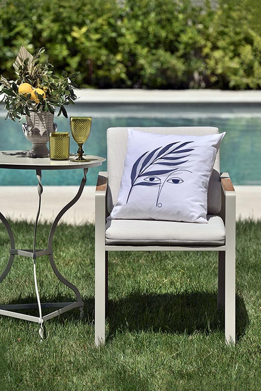 vigilia III Λευκό μαξιλάρι από 100% βαμβάκι, με ιδιαίτερο σχέδιο μπλε πρόσωπο και κλαδί ελιάς πάνω σε καρέκλα κήπου μπροστά από πισίνα.