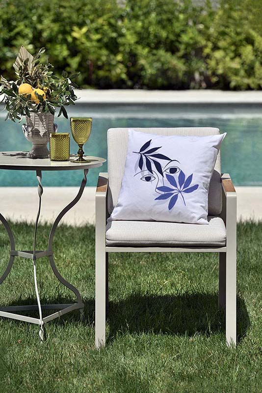 vigilia IV Λευκό μαξιλάρι από 100% βαμβάκι, με ιδιαίτερο σχέδιο μπλε πρόσωπο και κλαδί ελιάς πάνω σε καρέκλα κήπου μπροστά από πισίνα.