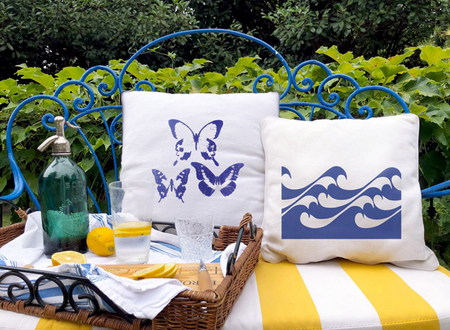 Τετράγωνα λευκά διακοσμητικά μαξιλάρια με μπλε σχέδια από πεταλούδες και κύματα, τοποθετημένα σε μπλε κλασικό μεταλλικό καναπεδάκι κήπου